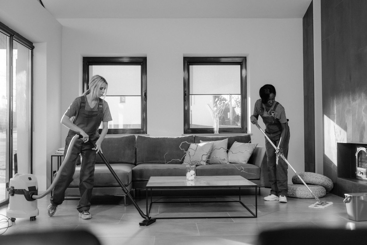 about-us Two women in work clothes cleaning a modern living room using a vacuum and mop.