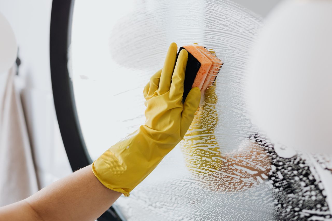 about-us Anonymous person in yellow rubber gloves using simple sponge for cleaning mirror in bathroom